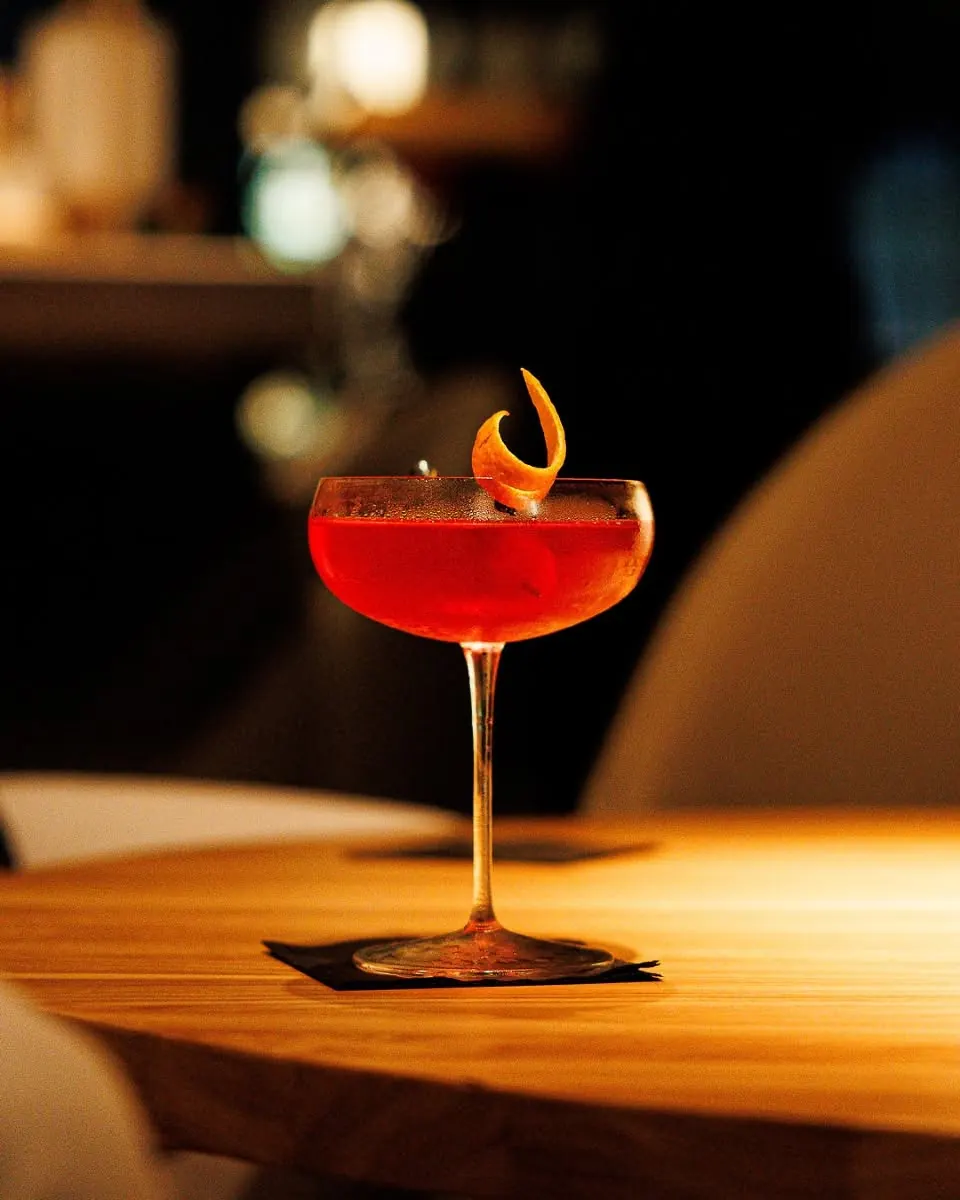 A red cocktail in a coupe glass on a lamplit table
