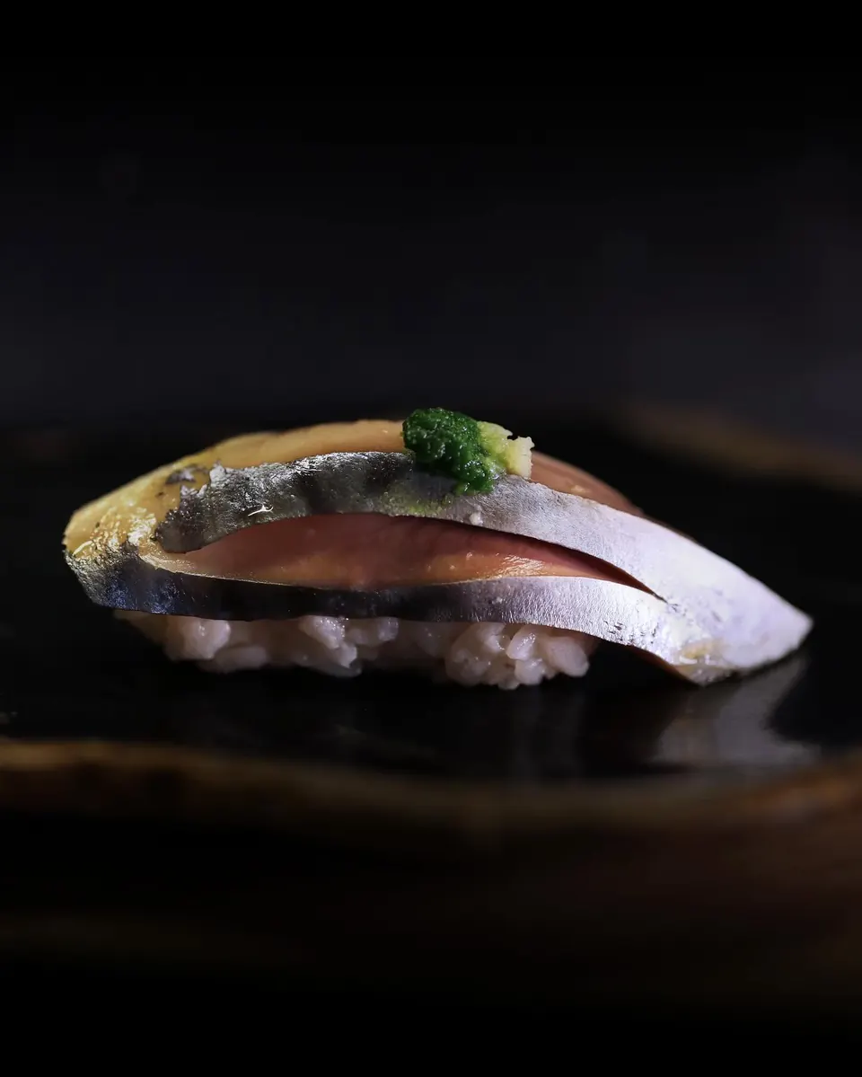 A single nigiri of silver-skinned fish on a dark plate